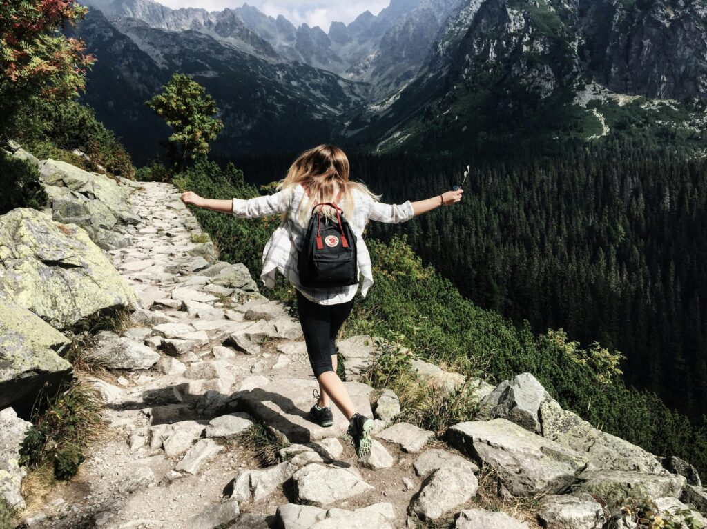 Woman walking across rocky trail in the mountains with arms raised up