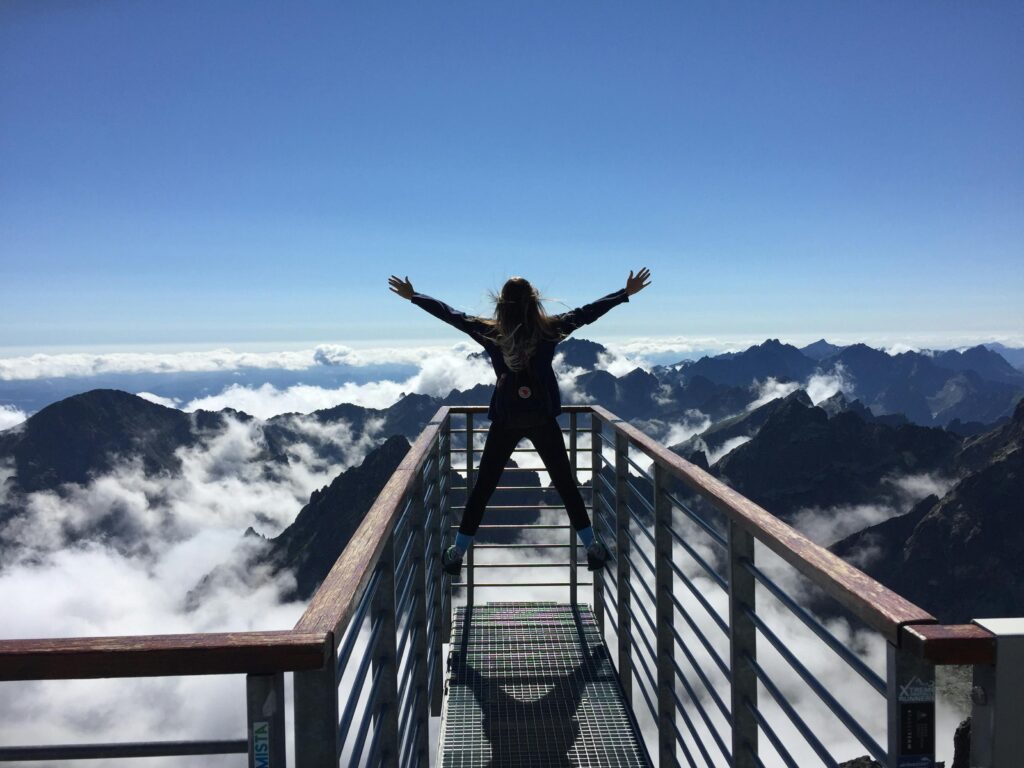 Woman on platform with arms in the air as she overlooks mountain peaks all around her
